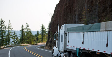 Big rig white American model bonnet semi truck tractor with plastic covered long bulk semi trailer driving on the curve mountain road with rock mountain cliff wall in Columbia River Gorge area