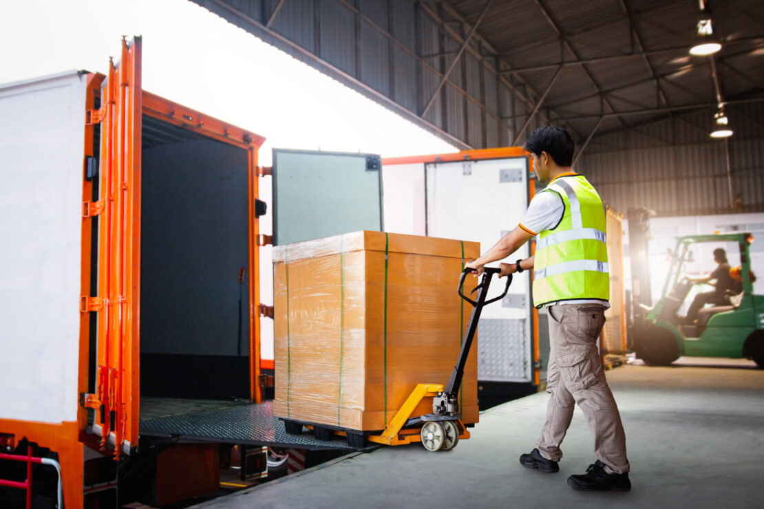 A man in a bright yellow vest is using a dolly to load a large, wooden box into a truck trailer.