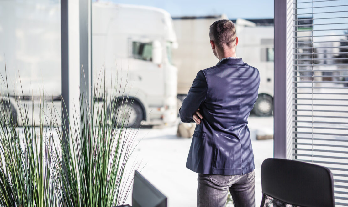 A man folds his arms and stares from his office out of a window, where there are several parked semi-trucks