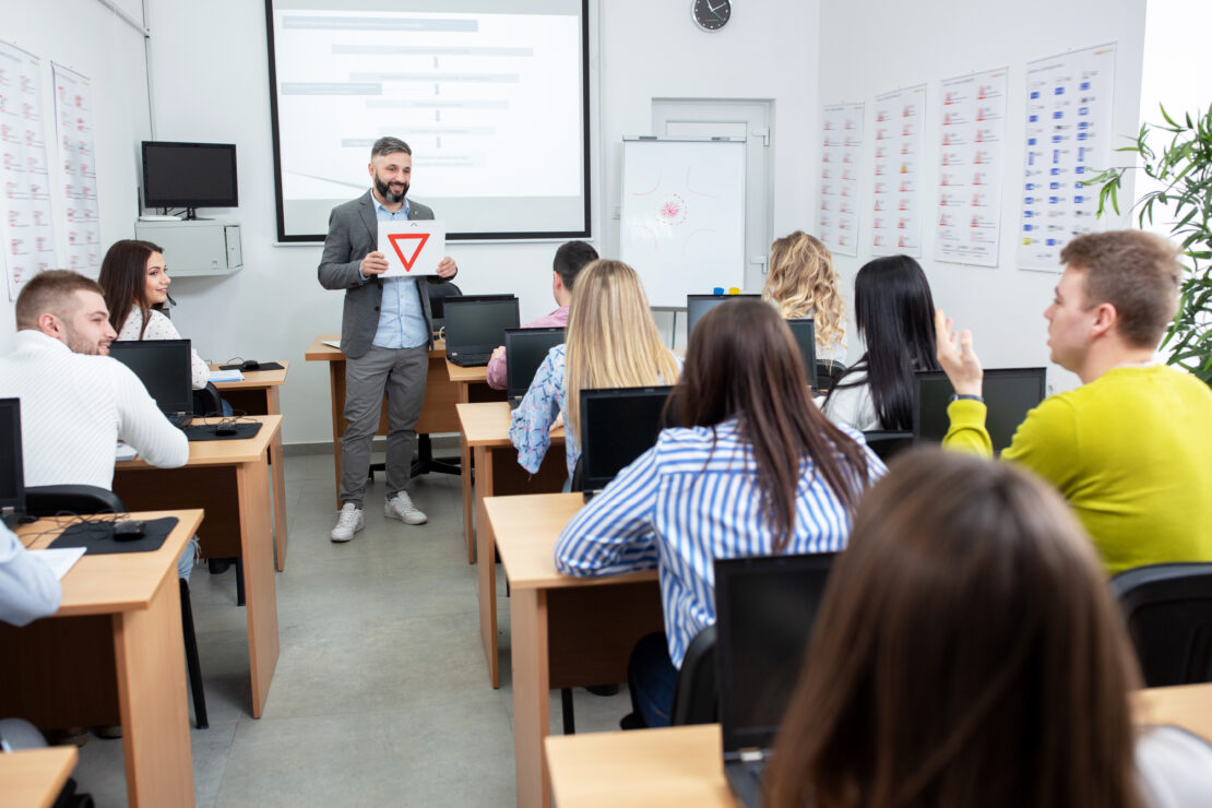 An instructor holds a sign with a red upside down triangle (sign for emergency) to a group of students who sit at desks with laptops.