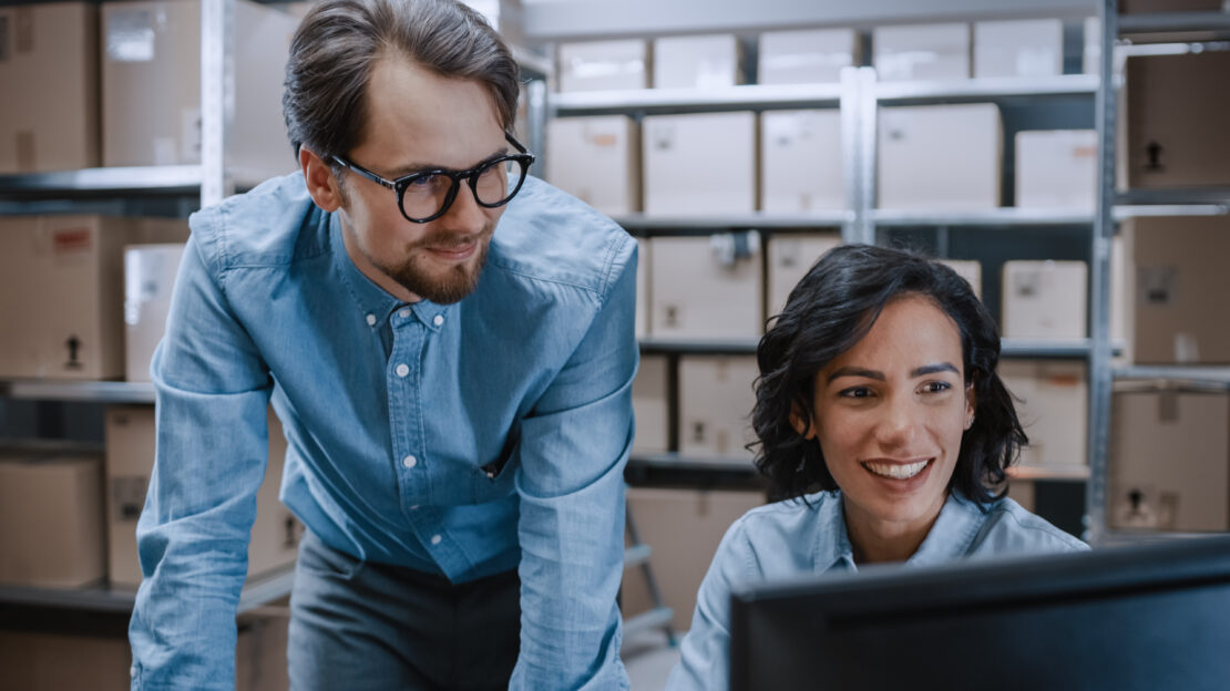 A man and woman wearing similar clothes are in a warehouse, smiling, and looking at a computer screen.