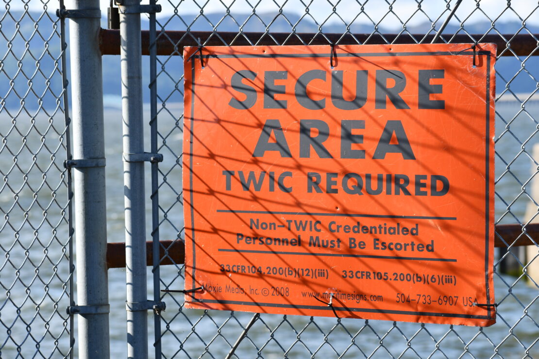 An orange sign zip tied to a fence that reads "Secure Area. TWIC Required. Non-TWIC Credentialed Personnel Must Be Escorted"
